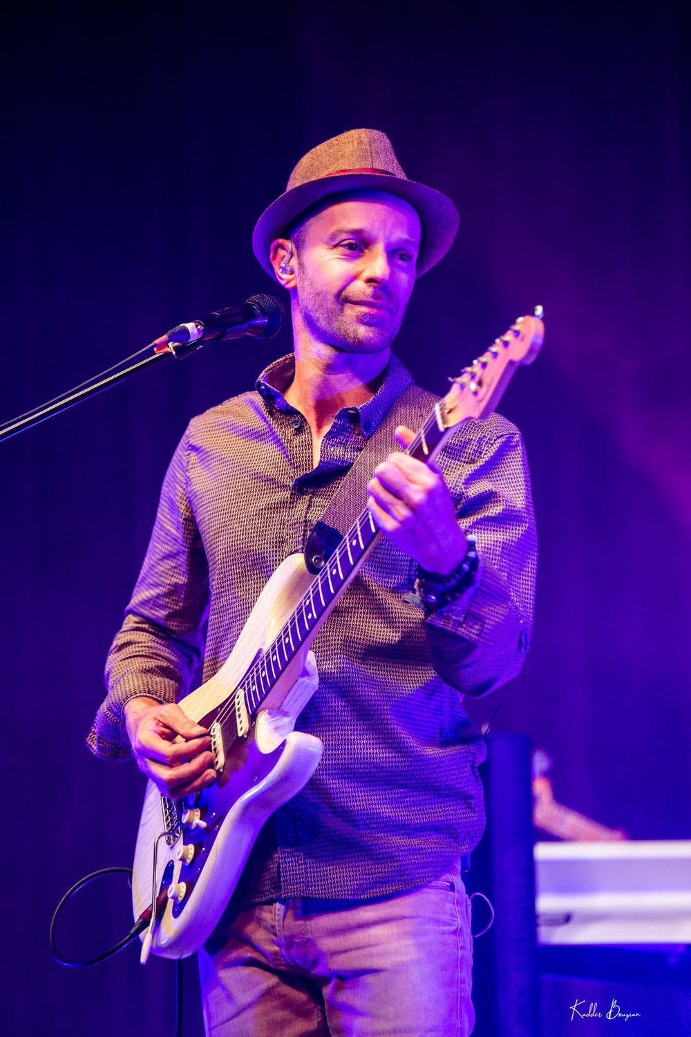 Cédric B. souriant sur scène, guitare électrique blanche
